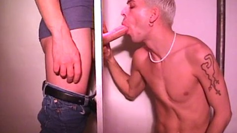 Le jeune blondinet s'amuse avec deux mecs taillés dans une cabine glory hole